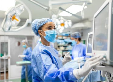 Anesthetist Working In Operating Theatre Wearing Protecive Gear checking monitors while sedating patient before surgical procedure in hospital