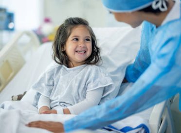 A female doctor of Indian decent checks in on a young female patient after her surgery.  The patient is sitting up in bed and appears happy.
